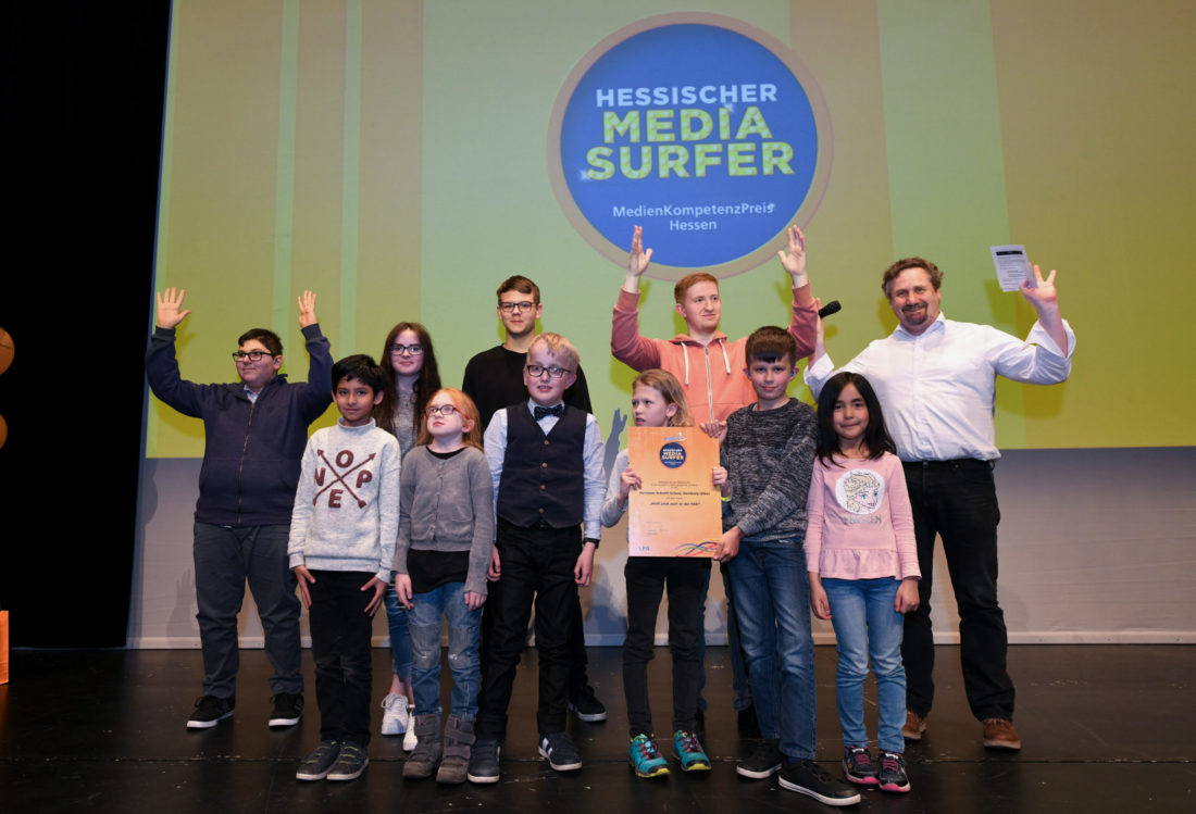 Eine Gruppe von Schülerinnen und Schülern hält die Hände nach oben und präsentiert eine Urkunde. Im Hintergrund steht an der Leinwand „Hessischer Mediasurfer"