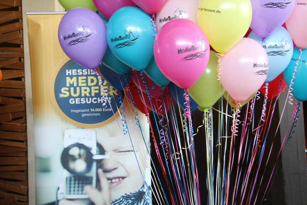 Viele bunte Luftballons. Im Hintergrund steht ein Rollup mit der Aufschrift: „Mediasurfer gesucht".