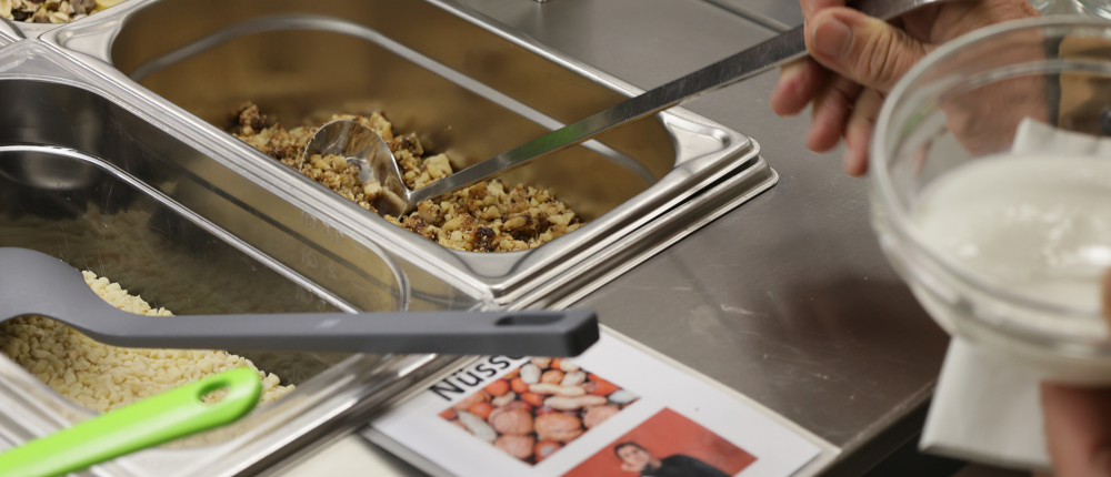 Müsli wird in einen Joghurt gefüllt. Darunter befindet sich ein Schild mit einem Bild von Nüssen und eine Person ist abgebildet, die in Gebärdensprache das Wort „Nüsse“ aufzeigt.