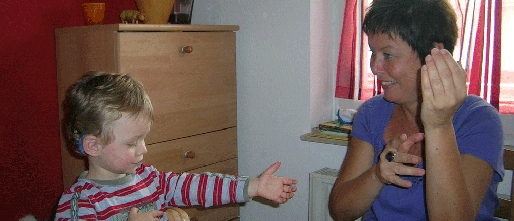 Frühförderung Ein kleines Kind mit Hörgerät steht vor einer Frau und reicht ihr die Hand. Die Frau scheint mit dem Kind in Gebärdensprache zu kommunizieren.