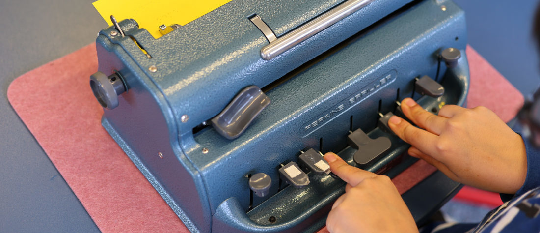 „Eine Punktschriftmaschine, an der ein Kind arbeitet. Die Maschine wird genutzt, um Texte in Braille-Schrift zu erstellen, um das Lesen und Schreiben für Menschen mit Sehbeeinträchtigungen zu ermöglichen.