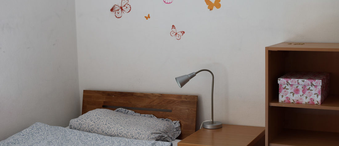 Ein Bett aus Holz mit einem Nachttisch und einer Lampe darauf. An der Wand befinden sich bunte Aufkleber.