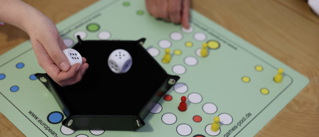 Ein „Mensch ärgere dich nicht“-Spiel, bei dem eine Hand mit einem großen Würfel würfelt. Die Zahlen auf dem Würfel sind als Erhebungen markiert, um das Spiel für Menschen mit Sehbeeinträchtigungen zugänglicher zu machen.