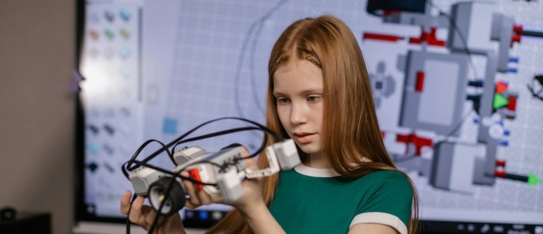 Ein Mädchen hält ein Objekt aus Klemmbausteinen in der Hand. Im Hintergrund ist ein Whiteboard mit digitalen Zeichnungen.