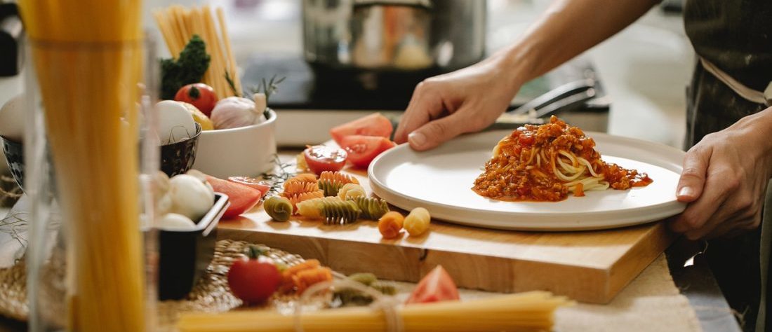 Eine Person hält einen Teller mit Spaghetti Bolognese. Daneben liegen getrocknete Nudeln und Hozbretter als Arbeitsfläche.