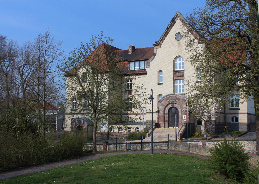 Blich auf das schöne große, alte Schulgebäude der Hermann-Schafft-Schule. Im Vordergrund eine Wiese und grüne Bäume