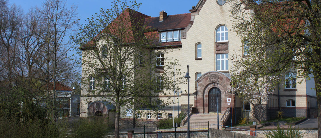 Blich auf das schöne große, alte Schulgebäude der Hermann-Schafft-Schule. Im Vordergrund eine Wiese und grüne Bäume