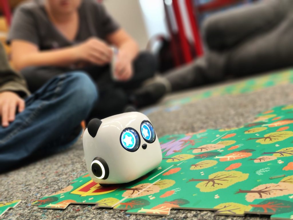 Ein kleiner Roboter Panda, der über ein Spielfeld rollt. Im Hintergrund sitzen Kinder, die zuschauen.