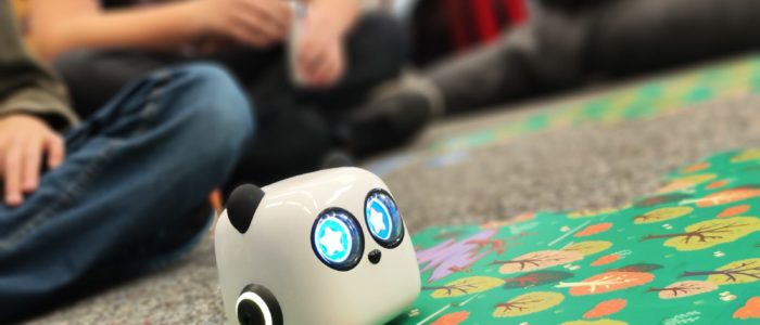 MakerSpace Panda Ein kleiner Roboter Panda, der über ein Spielfeld rollt. Im Hintergrund sitzen Kinder, die zuschauen.