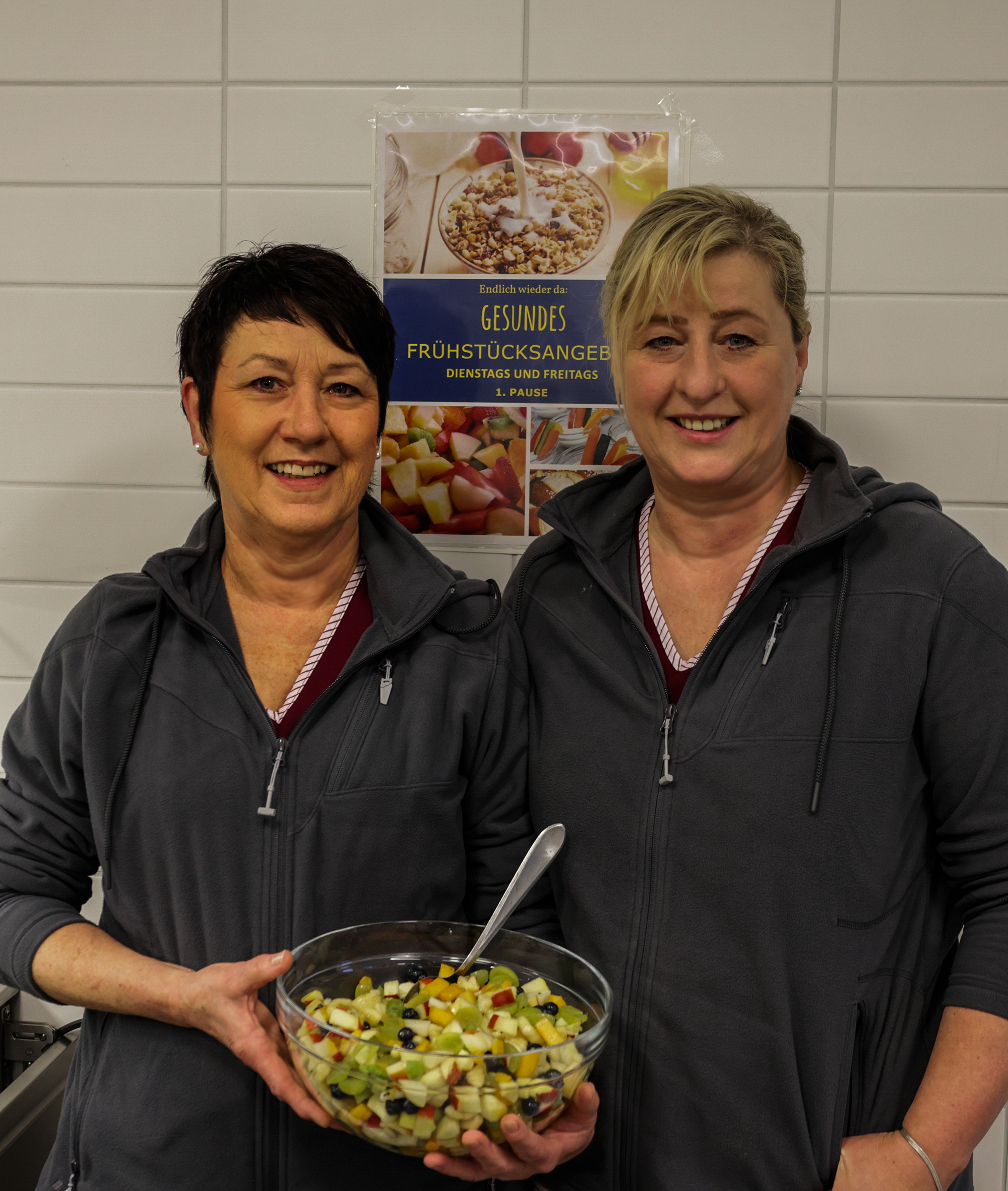 Das Mensa Team schaut freundlich in die Kamera. In der Hand halten Sie eine Schüssel mit Obstsalat. Im Hintergrund sieht man ein Plakat mit dem Frühstücksangebot der Schule.