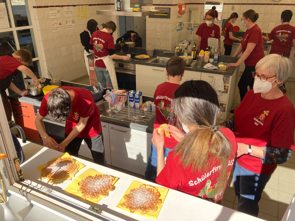 Im Vordergrund Waffeln. Im Hintergrund viele Schülerinnen und Schüler, die fleißig Waffeln backen.